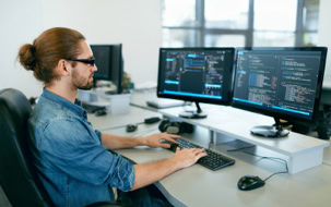 Mann mit Brille arbeitet an einem Schreibtisch mit zwei Monitoren, auf denen Code angezeigt wird.