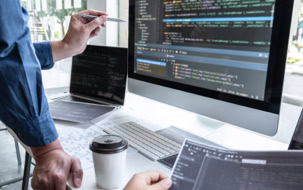 Zwei Personen arbeiten an Computern mit Code auf den Bildschirmen, eine Person zeigt mit einem Stift auf den Monitor, während die andere ein Dokument hält; ein Kaffeebecher steht auf dem Tisch.